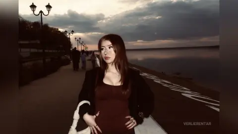 Young woman in a dark casual outfit standing by a lake at sunset under a moody, cloudy sky.
