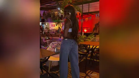 Young woman in a white crop top and baggy jeans posing at a vibrant outdoor rooftop lounge at night.