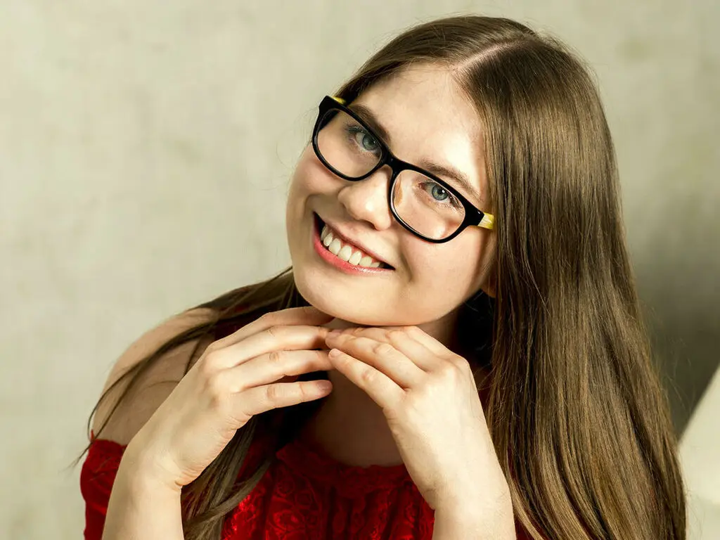 Smiling young woman with long brown hair and black glasses wearing a red lace top in a bright indoor setting.