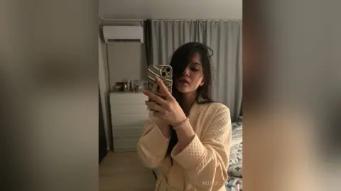 Young woman in a cream quilted robe taking a mirror selfie in a cozy bedroom setting.