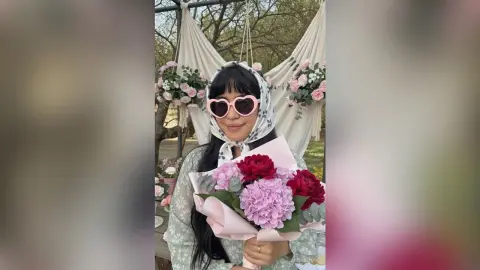 Smiling woman in a white headscarf holding a large pink hydrangea bouquet in a dreamy garden setting.