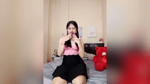Cheerful young woman in a pink lace top and black skirt sitting on a bed next to a red teddy bear.