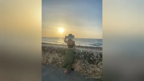 Woman in olive green outfit and sun hat gazing at the ocean sunset from a stone pier.
