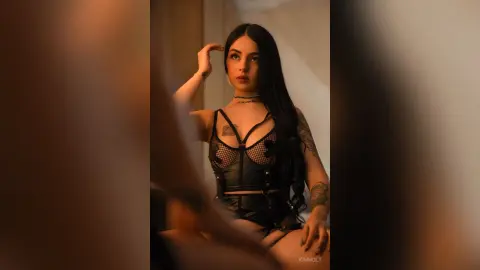 Young woman with long dark hair wearing a black lace corset outfit in a moody, dimly lit indoor setting.