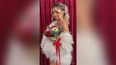 Glamorous woman in a white feathered cocktail dress holding a colorful floral bouquet against red velvet curtains.