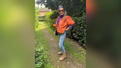 Smiling woman in orange blouse and purple scarf standing on a garden path near lush green bushes.