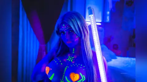 AndreaKalo poses with long blonde hair and neon green body paint on her chest and face under bright blue UV lighting.