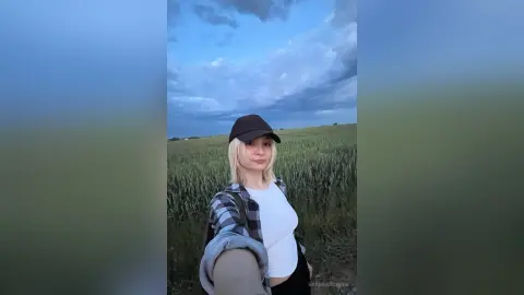 BarbaraGreen wears a black cap and a plaid shirt over a white tank top while standing in a green field under a cloudy sky.