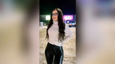 Stylish woman in a pink turtleneck sweater and black leather pants posing outdoors at night.