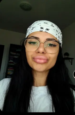 Smiling young woman with long dark hair, glasses, and a white patterned headband in a bright indoor setting.
