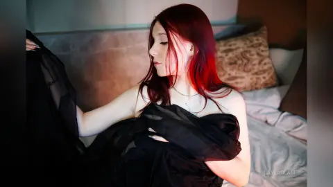 Red-haired woman in a black silk robe relaxing on a bed in a moody, dimly lit bedroom setting.