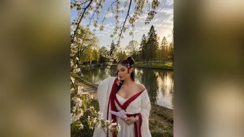 IngridSaint poses in a white and red dress by a river under blooming trees at sunset.