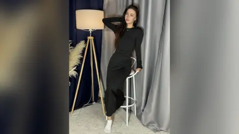 Young woman in a long black dress posing elegantly by a lamp in a minimalist studio setting.