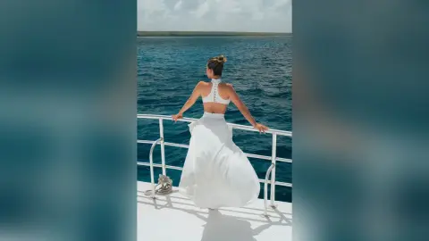 Elegant woman in a white halter crop top and flowing skirt standing on a boat deck overlooking the blue ocean.