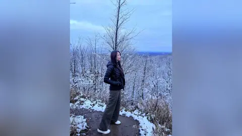 Woman in a black puffer jacket and grey trousers standing in a snowy winter forest under a cloudy sky.