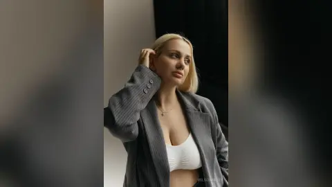 Blonde woman in a grey pinstripe blazer and white crop top posing thoughtfully in a minimalist indoor setting.