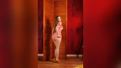 Young woman in a red floral mini dress posing against a wooden screen in a moody red studio setting.