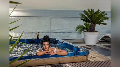MissyJolie leans against the edge of a blue hot tub overlooking the ocean, wearing white bikini tops or lingerie.