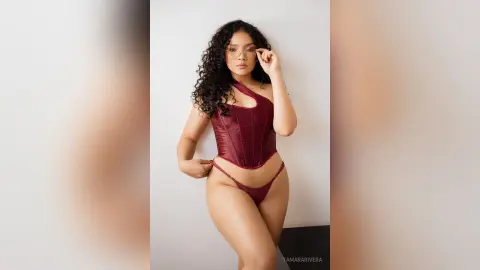 Curvy woman with curly hair wearing a burgundy satin corset and matching lingerie in a minimalist studio setting.