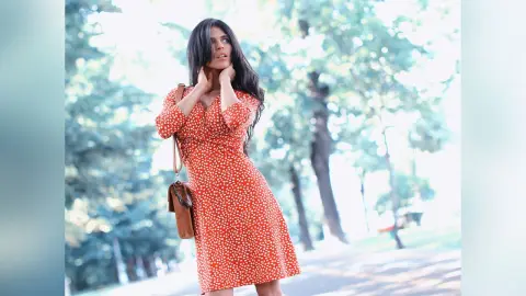 Young woman in a red polka dot dress standing in a sunny park with a brown leather shoulder bag.