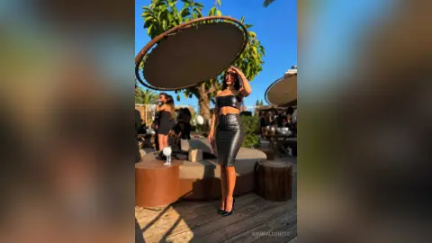 BiankaLeoness poses outdoors in a black crop top and tight black leather skirt, looking away under the sun.