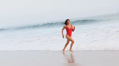 Cheerful woman in a red one-piece swimsuit running along a sunny beach with ocean waves.