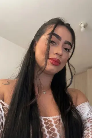 Young woman with long dark hair and red lipstick wearing a white lace top in a bright indoor setting.