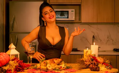 Smiling woman in a black dress serving a roasted turkey dinner at a festive Thanksgiving meal in a kitchen.