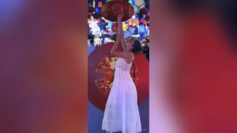 AnaMathers stands outdoors at night wearing a white sundress and reaching her arms up toward a large red lantern.