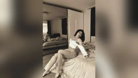 Elegant woman in a white off-the-shoulder top sitting on a bed in a bright, minimalist bedroom.