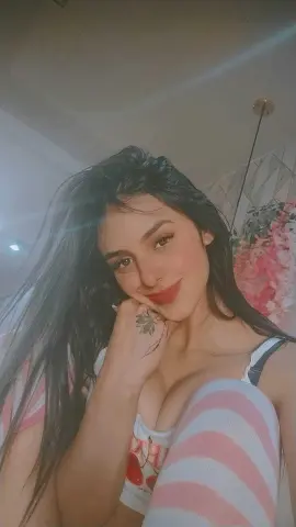 Smiling young woman with long dark hair wearing a white and pink striped tank top in a soft indoor setting.