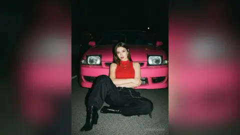 Stylish woman in a red tank top and black trousers sitting in front of a pink car at night.