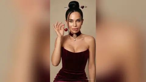 Glamorous woman in a burgundy velvet dress holding a cherry against a neutral studio background.