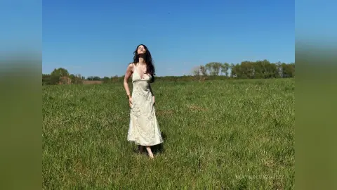 Serene woman in a flowing white lace maxi dress standing in a sunny green meadow under a clear blue sky.