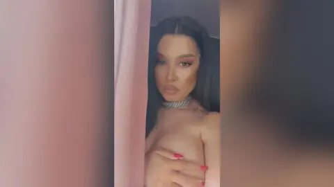 AnnaBanks poses with long dark hair and a silver choker, holding her bare breasts with her hands against a soft pink background.