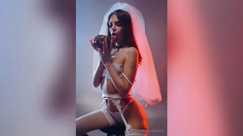 Sultry woman in white lace lingerie and bridal veil holding a dark fruit in moody studio lighting.