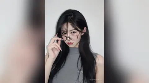 Young woman with long black hair wearing a grey turtleneck holding glasses in a minimalist indoor setting.