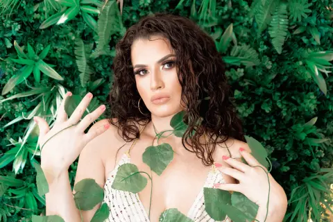 Beautiful woman with wavy hair in a white sequin bikini posing against a lush green tropical leaf background.