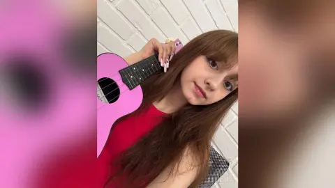 Young woman with long brown hair holding a pink ukulele against a white brick wall in a relaxed mood.