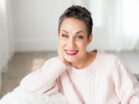 Smiling woman with short pixie hair wearing a cozy pink knit sweater in a bright, airy room.