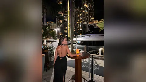 EvelynMills stands with her back turned, wearing a black backless dress that exposes her bare back and skin.