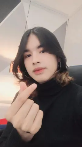 Young woman in a black turtleneck sweater making a finger heart gesture in a cozy indoor setting.