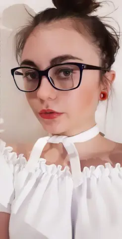 Young woman with glasses and red lipstick wearing a white off-the-shoulder blouse in a bright indoor setting.