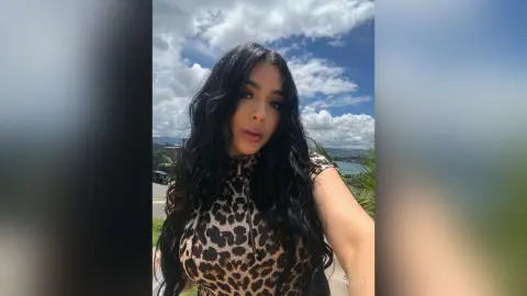 IvyStuart poses outdoors with long, wavy black hair, wearing a tight leopard print top against a cloudy blue sky.
