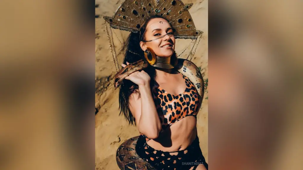 ShanitaDiaz smiles while posing with one arm raised, wearing a red lace bra and gold earrings against a soft indoor background.
