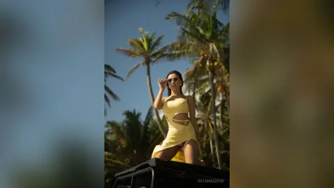 Stylish woman in a yellow summer cutout dress posing under palm trees in a sunny tropical setting.