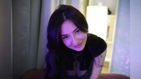 Smiling young woman in a black outfit posing in a cozy room with soft purple ambient lighting.