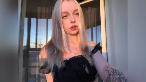 Blonde woman in a black ruffled top posing by a window during golden hour sunset.