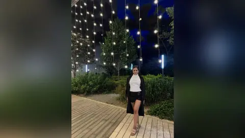 Stylish woman in a white turtleneck and black skirt standing on a wooden walkway under festive string lights.