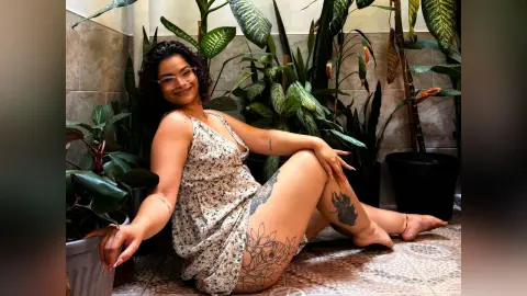 Smiling woman in a floral sundress sitting amidst lush green houseplants in a bright indoor garden setting.